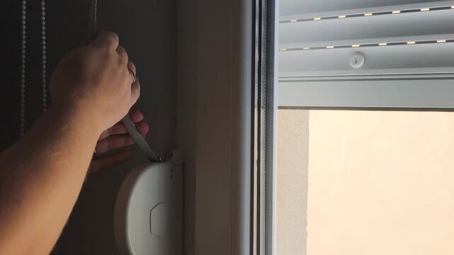 Lowering outdoor roller shutter with manual strap cord protects room from sun. White roller shutter closes slowly on glass window to create shade, regulate temperature, and ensure home privacy.