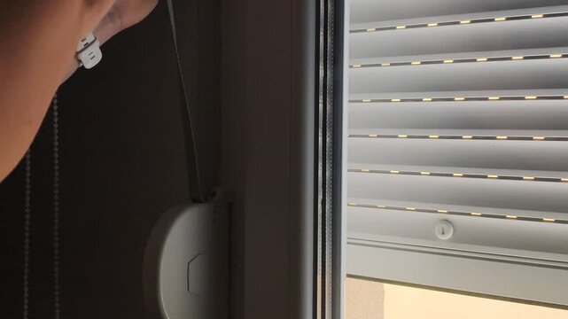 Hands pull belt cord to open window shutters in home interior. White window shutters rise slowly on glass frame to let bright daylight inside, illuminate room space, and offer nice outdoor view.