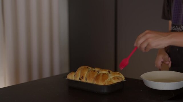 Hand brushing a shiny glaze onto a golden, freshly baked sweet bread loaf with raisins in a baking pan on a dark kitchen counter