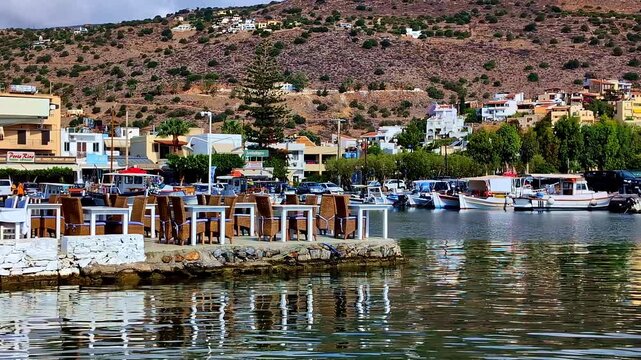 Small boats line a waterfront harbor beneath a dry hillside with clustered homes, Crete, Greece
