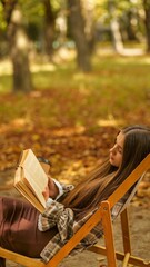 Young woman relaxing in a chair reading a book in a park during autumn