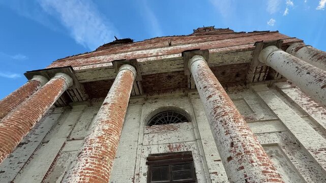 The facade of an old building in the classical style, featuring massive columns, dilapidated brickwork, and an arched window against a backdrop of a blue sky.