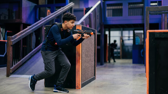 Teenage boy playing laser tag in an indoor arena. Young male holding a blaster vest crouching behind an obstacle during game activity