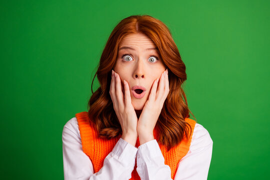Surprised redhead woman with hands on cheeks expressing shock and astonishment, wide eyes and open mouth against a vibrant green background