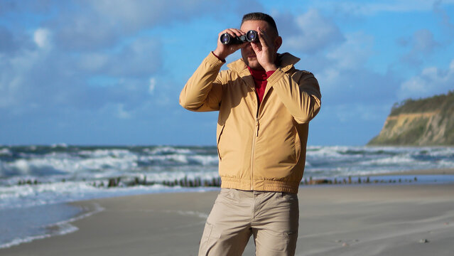Front view of a man holding field binoculars while standing on the windy sea coast in autumn.