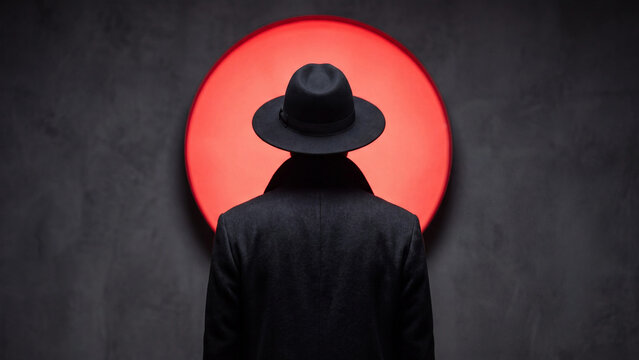 Detective wearing fedora hat and dark coat standing in front of bright red circular light.