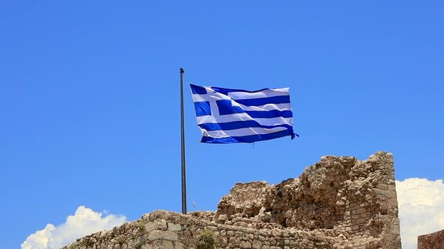 A Greek flag waving against a clear blue sky. The flag features blue and white stripes with a cross in the upper left corner. Rocky terrain is visible in the background.