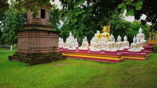 Row of white and gold Buddha statues seated beside an old brick pagoda in a traditional Thai temple.