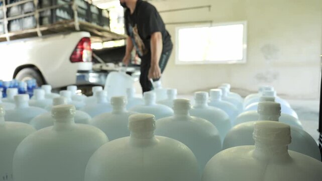 Rows of drinking water gallon bottles at a water bottling factory, with a worker in the background loading gallons onto a pickup truck for delivery and distribution.	