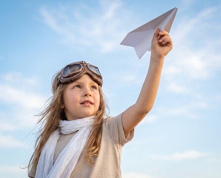 Little child playing with a white paper airplane wearing vintage pilot goggles and scarf