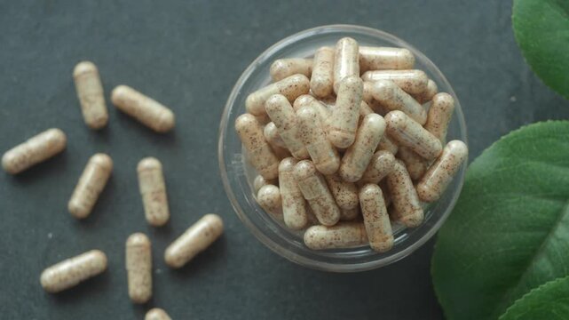 Herbal supplement capsules in glass bowl with green leaves on dark background top view cinematic slow motion motion