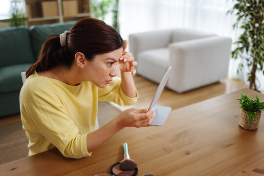 Young woman closely inspecting her forehead skin in a small mirror, focusing on wrinkles or imperfections during her daily beauty and skincare routine at home