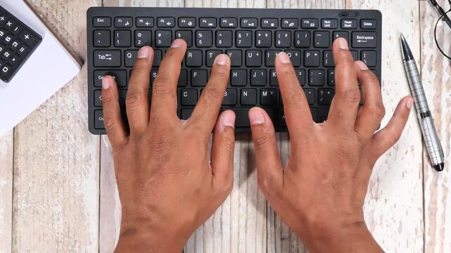 Hands typing on black wireless keyboard over light wooden desk with office supplies