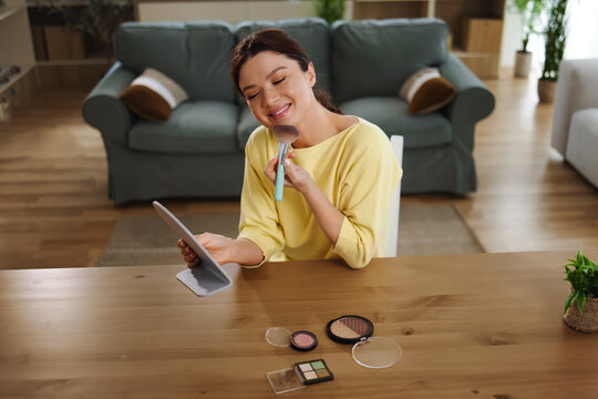 Young woman applying face powder with a brush, looking at a mirror and smiling, doing her morning makeup routine at home for beauty and self-care