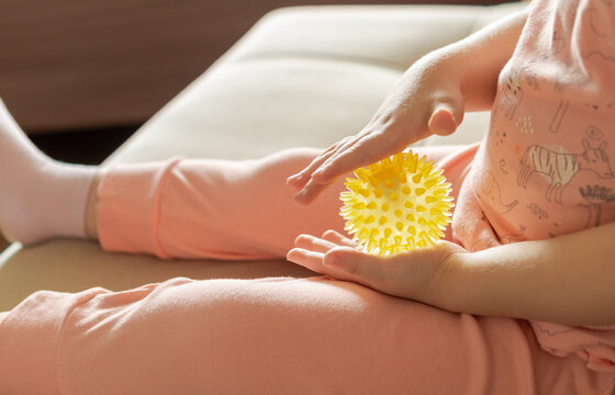 Unrecognizable child exploring a yellow spiky sensory massage ball at home. Tactile stimulation, sensory development, hand coordination and fine motor skills concept.