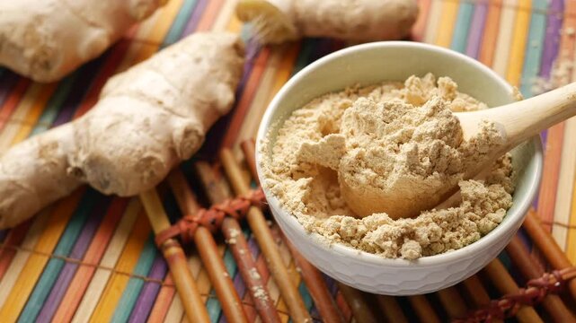 Wooden spoon lifting organic ginger powder from white bowl on colorful bamboo mat with fresh ginger roots
