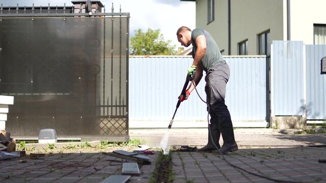 Man using a pressure washer to blast away dirt and clean a driveway trench drain with a static camera shot.