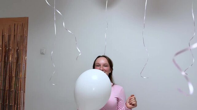 A carefree young woman in a pink top playfully interacts with white balloons, pulling them from the ceiling against a bright wall in a cheerful indoor setting.