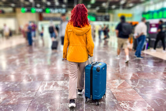Back view of a solo female traveler pulling a turquoise suitcase through a busy, blurred airport terminal hall, setting off on an international journey, vacation, flight departure, or travel trip
