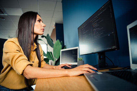 Female software developer coding at dual-monitor workstation in modern office, focused programmer working on laptop and desktop with code on screen