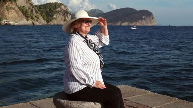 Mature Woman Relaxing by the Sea on Vacation. Mature woman in sun hat and sunglasses sitting by the sea and waving her hand, with coastal mountains and a boat in the background during summer vacation.