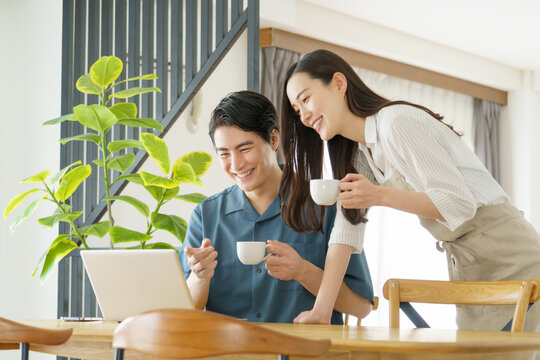 自宅のテーブルでノートパソコンで作業をする男性とマグカップを持つ女性の若い日本人パワーカップル