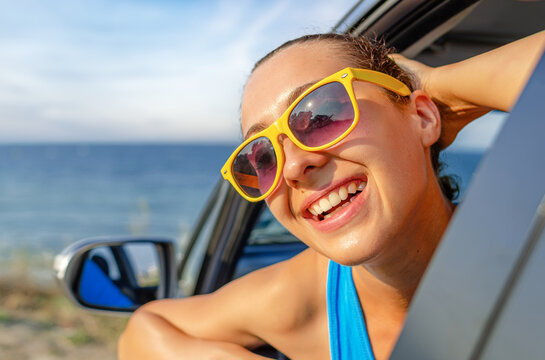 A cheerful young woman in yellow sunglasses leans out of a car window by the sea during a summer road trip.