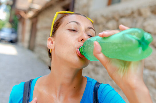 A thirsty young woman drinks fresh water directly from a green plastic bottle on a hot sunny day outdoors.