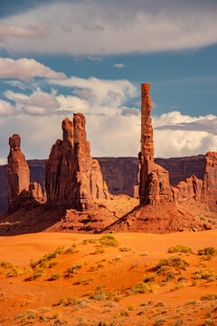 View of the iconic sandstone buttes and the Totem Pole spire in the desert landscape under a cloudy blue sky in Oljato-Monument Valley, Arizona, United States.