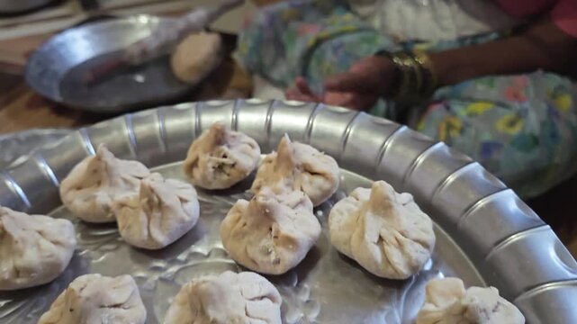 Close up details of uncooked ukdiche modak arranged on a traditional silver plate as a woman sits on the floor carefully shaping and stuffing the sweet dumplings for the Maharashtrian Ganesh festival.