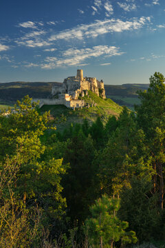View of the historic Spis Castle perched on a grassy hill surrounded by lush pine forests under a clear blue sky during the golden hour in Spisske Podhradie, Kosicky kraj, Slovakia.