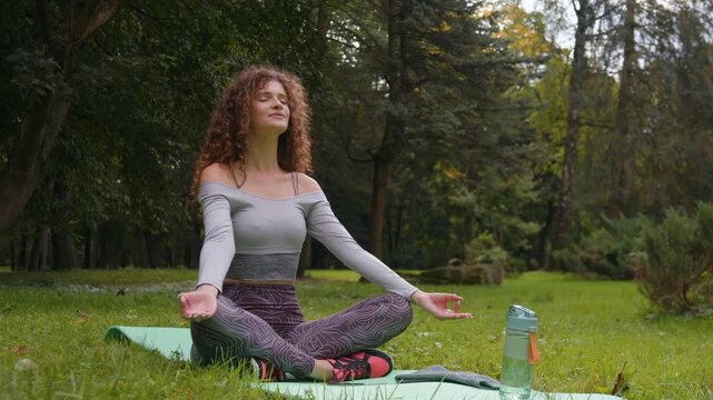 Calm Caucasian woman yogi girl sitting in lotus position relaxing practising meditation breathing exercise in park at summer. Female with closed eyes meditating doing yoga mindfulness workout outdoors