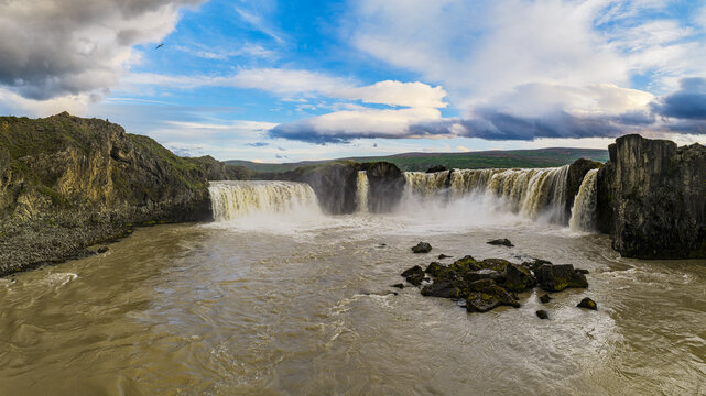 View of the powerful Godafoss Waterfall cascading over rugged basalt cliffs into a turbulent river under a dramatic cloudy sky in Thingeyjarsveit, Iceland.