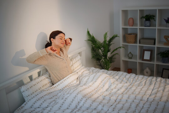 Woman waking up in the morning feeling refreshed and well-rested, stretching arms in bed, enjoying comfort and relaxation in her bright bedroom at home