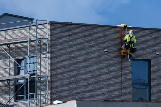 View of a construction worker in high-visibility gear and safety harness working on the exterior brick wall of a building with scaffolding nearby in Vilnius, Vilnius County, Lithuania.