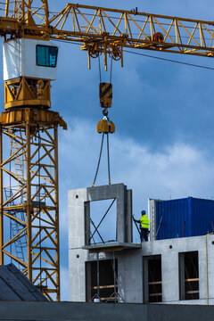 View of a construction site with a yellow tower crane lifting a concrete panel and a worker in a high-visibility jacket standing on the structure in Vilnius, Vilnius County, Lithuania.