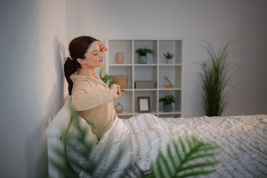 Young woman waking up in her comfortable bed, stretching with her eyes closed, feeling the warm morning sunlight on her face, embracing comfort and relaxation at home