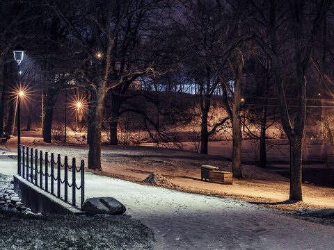 View of a snow-covered park path at night illuminated by glowing street lamps, with bare trees and a metal railing along a small bridge in Latvija.