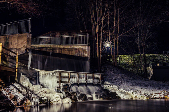 View of a frozen waterfall and dam at night with icicles hanging from wooden railings, bare trees, and a bright starburst light reflection in Latvia.