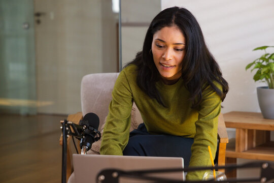 African American woman in green sweater leaning in armchair at studio, recording on laptop with mic