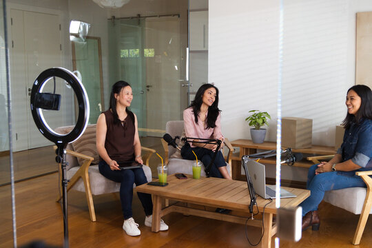 Diverse female colleagues in casual clothing recording podcast in studio with ring light, mics