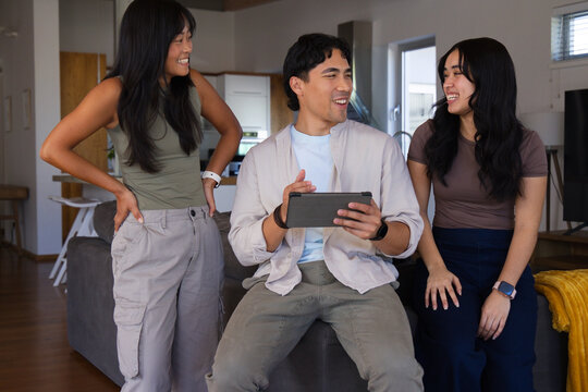 Diverse friends laughing and chatting on gray sofa in modern home while holding tablet
