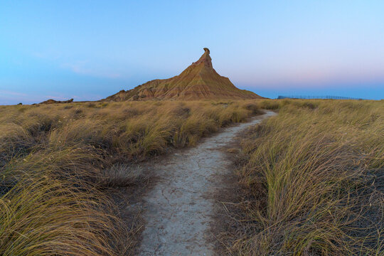 View of the Castildetierra rock formation in the Bardenas Reales desert with a dirt path winding through dry grasses under a clear sky in Spain.