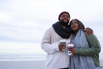Obraz premium Couple standing close on beach holding cups, wearing knit sweaters, scarves and beanie, copy space