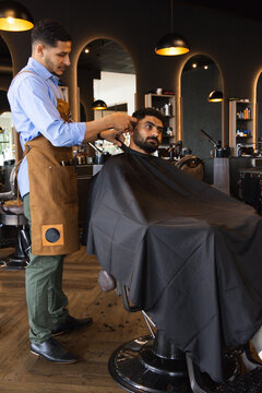 African American barber and client trimming hair and beard at barbershop apron, scissors