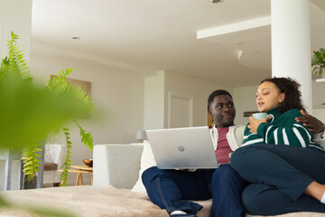 Obraz premium African American couple sitting on sofa in living room, holding laptop, cradling mug, copy space