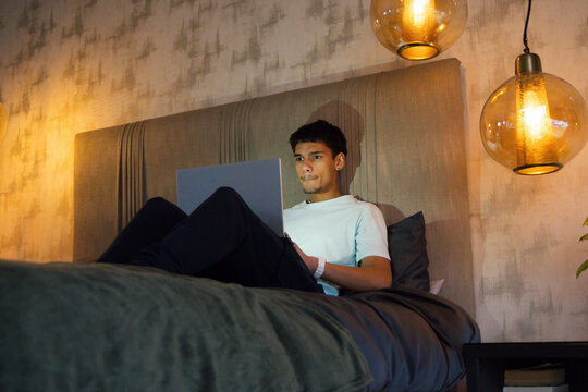 African American man sitting on bed using laptop with padded headboard under amber pendant lamps