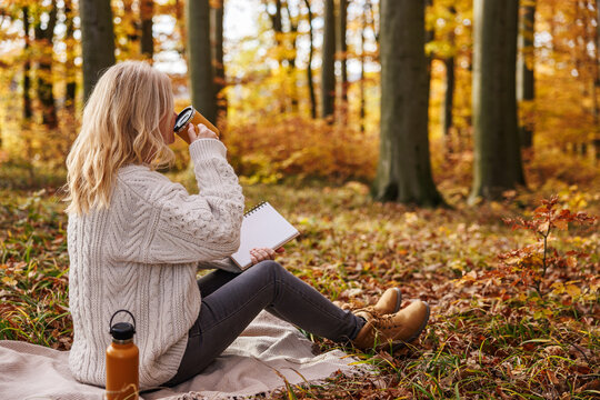 Woman relaxing in autumn forest while writing diary and drinking hot drink. Journaling offline for good mental wellbeing and mindfulness intentions
