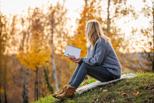 Woman practicing mindfulness and slow living through journaling. Relaxed woman is writing her diary and resting in autumn park