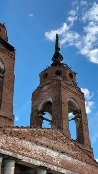 A fragment of a half‑ruined brick bell tower with arched openings, dilapidated masonry, and a dark spire against a bright blue sky with light, feathery clouds.